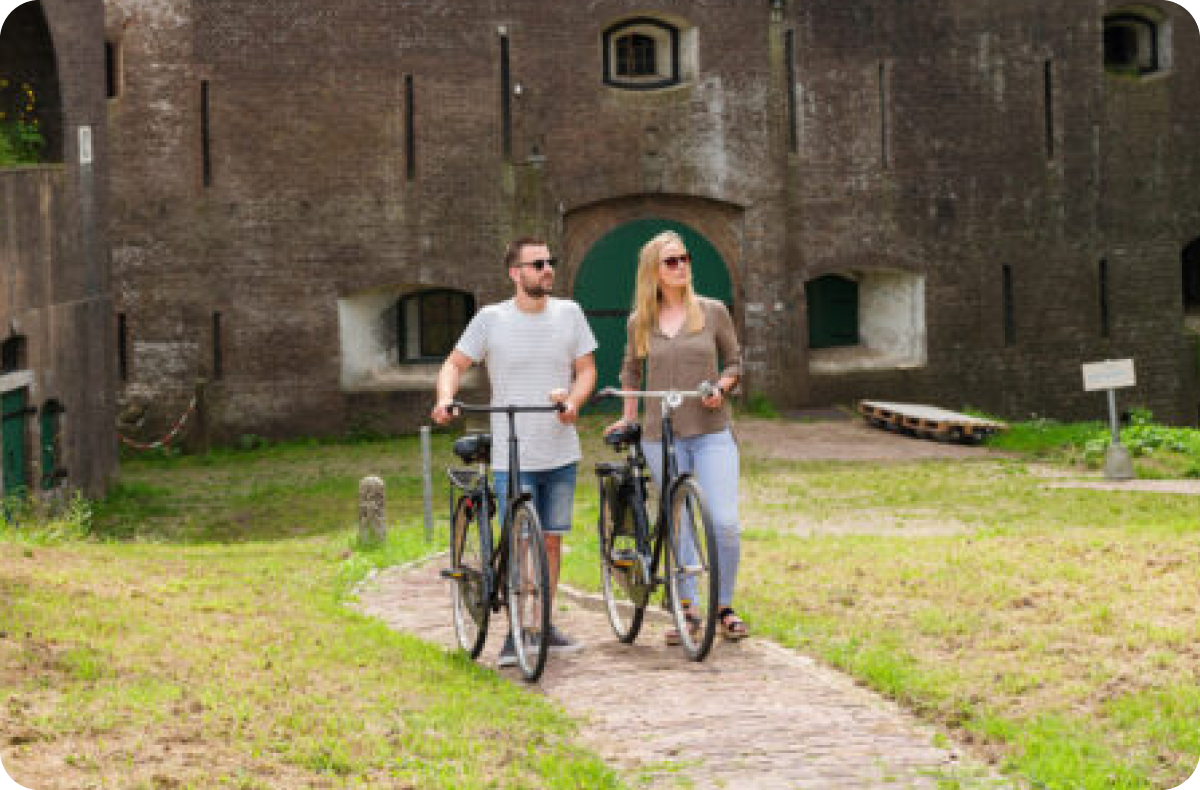 Een man en een vrouw lopen met hun fietsen in de hand over een pad voor een fort. Er is veel groen om hen heen.