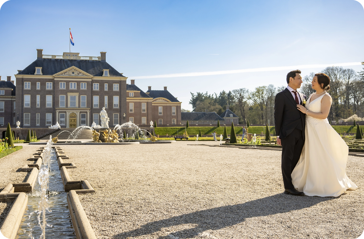Een bruidspaar staat in de tuin van Paleis Het Loo. Op de achtergrond zie je het paleis, een fontein en bomen.