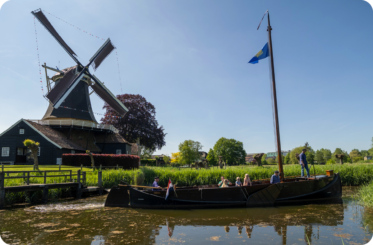 Een schuit met mensen vaart langs een windmolen, een groene oever en over glad water.