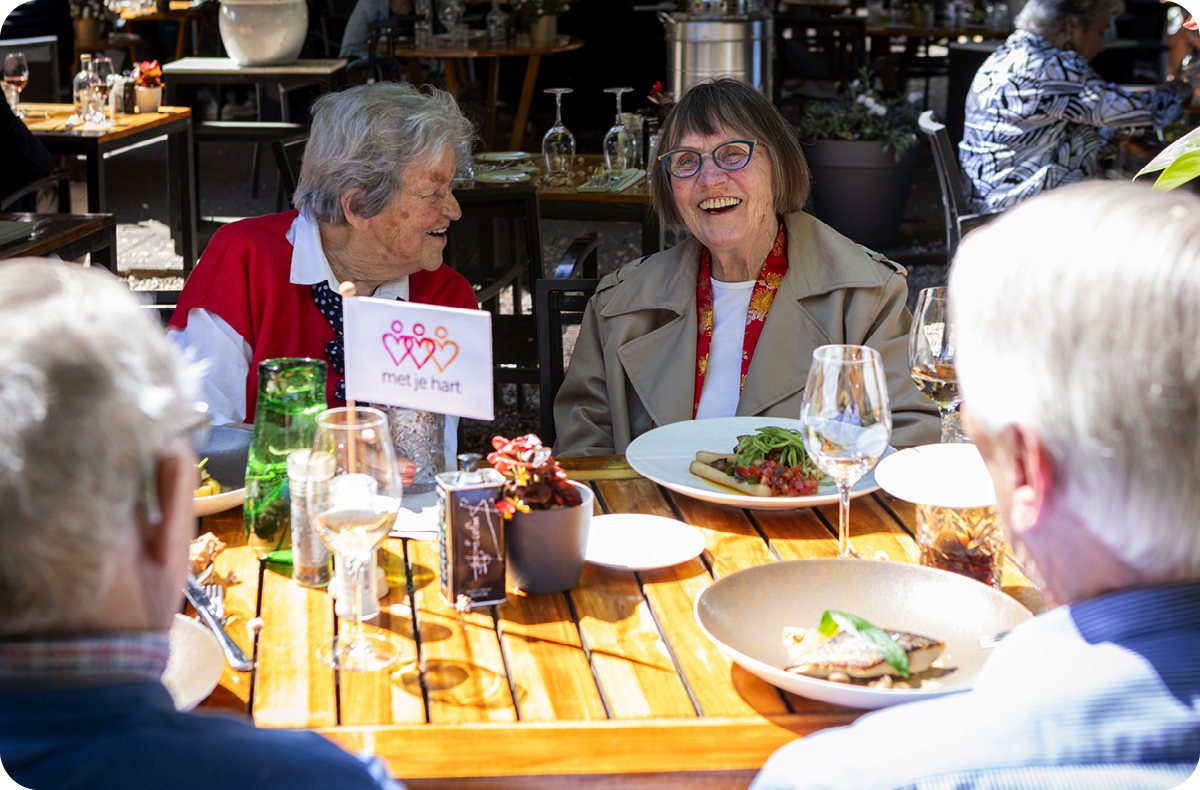 Ouderen praten en lachen met elkaar. Op tafel staan borden, glazen en een vlaggetje van Stichting met je hart.