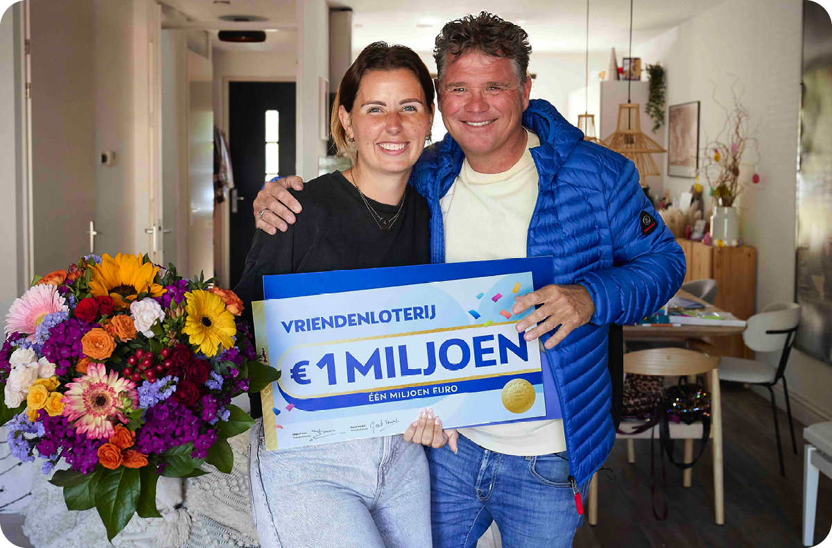 VriendenLoterij-ambassadeur Wolter Kroes staat met een lachende vrouw in de woonkamer en ze houden samen een grote loterijcheque van €1 miljoen vast. De vrouw heeft ook een kleurrijk boeket bloemen in haar hand.