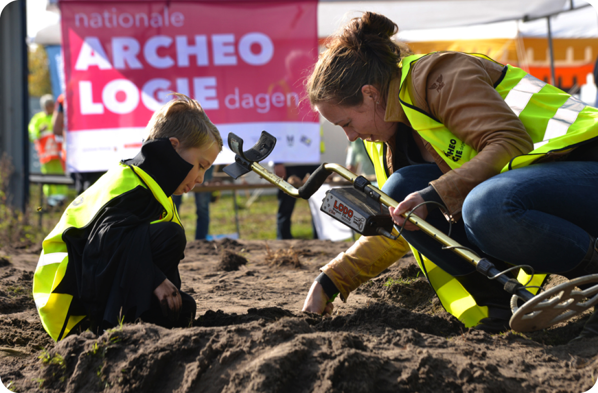 Een vrouw en kind in gele hesjes graven tijdens de Nationale Archeologiedagen. Achter hen hangt een vlag ervan.