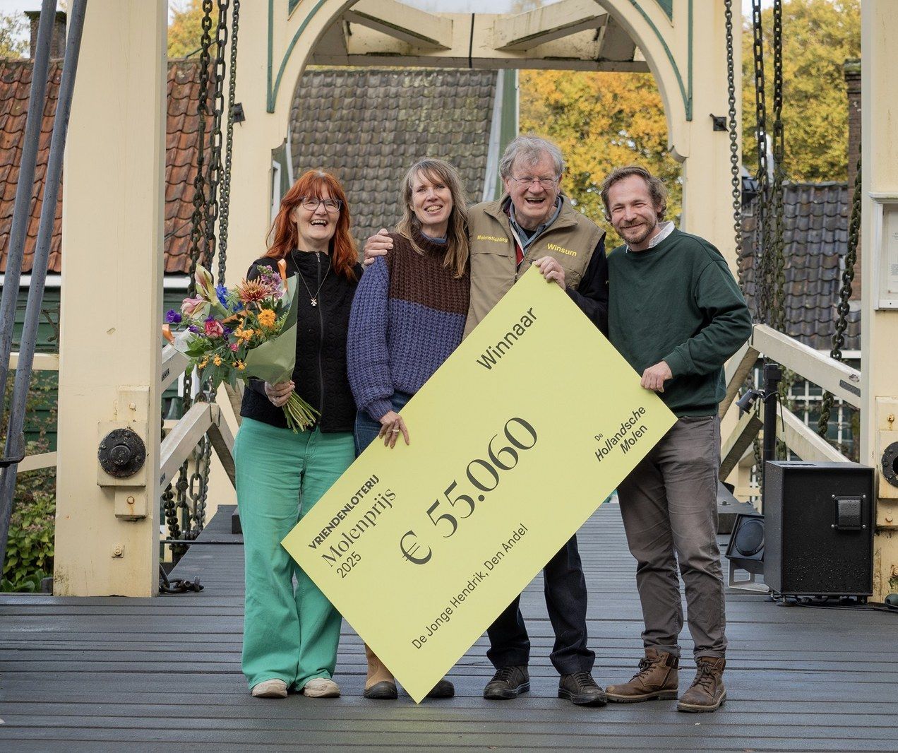 Vier mensen staan op een brug en houden een grote cheque van de VriendenLoterij Molenprijs vast ter waarde van €55.060. Eén persoon heeft een bos bloemen.