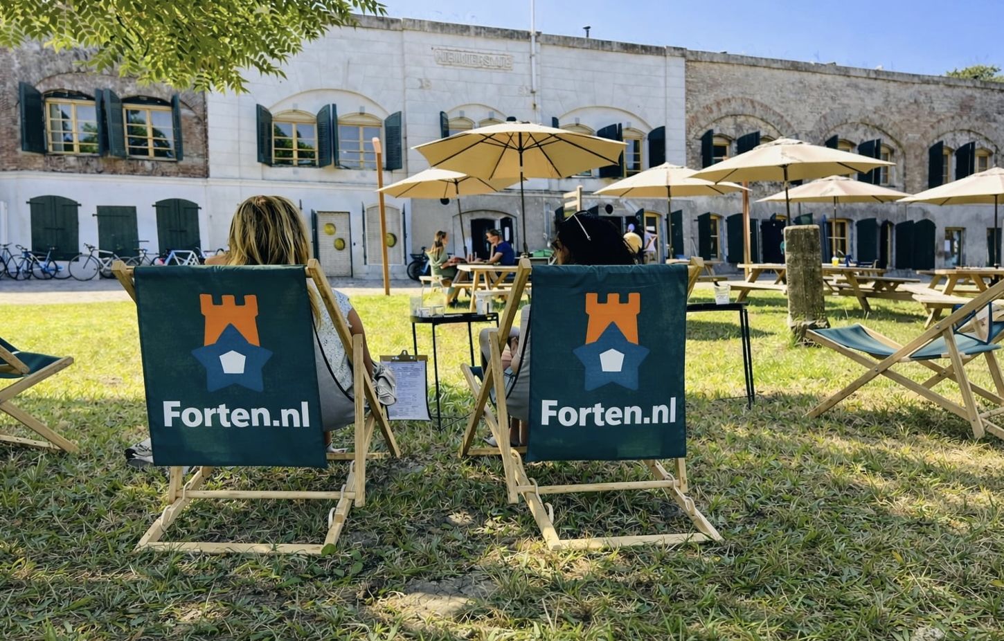 Twee mensen zitten op strandstoelen van “Forten.nl” op het gras. Op de achtergrond zie je een stenen fort, parasols, tafels en een zonnige lucht.