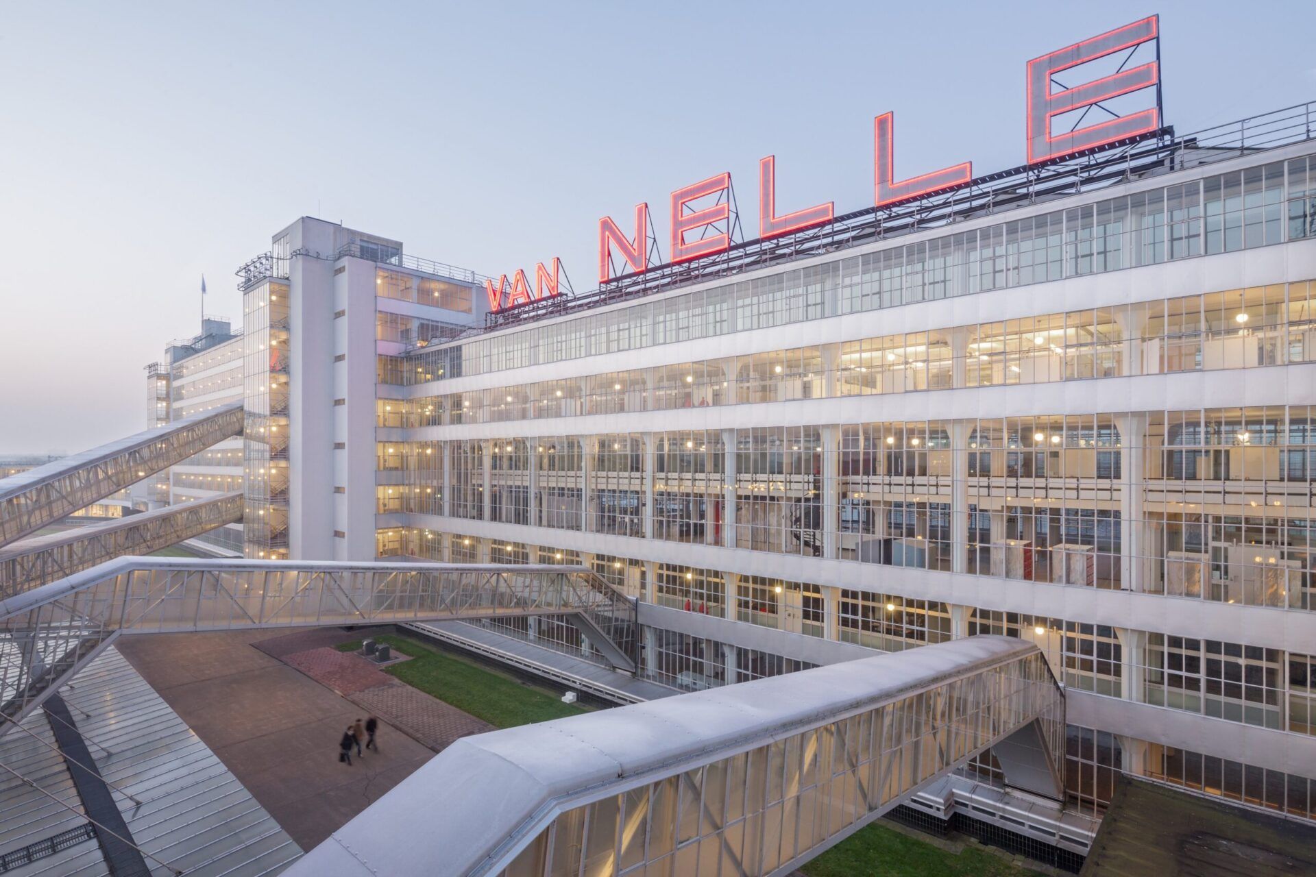 Buitenkant van een grote industrieel gebouw met veel glas, witte verdiepingslagen en loopbruggen aan de buitenkant. Op het dak staat in grote rode letters Van Nelle.