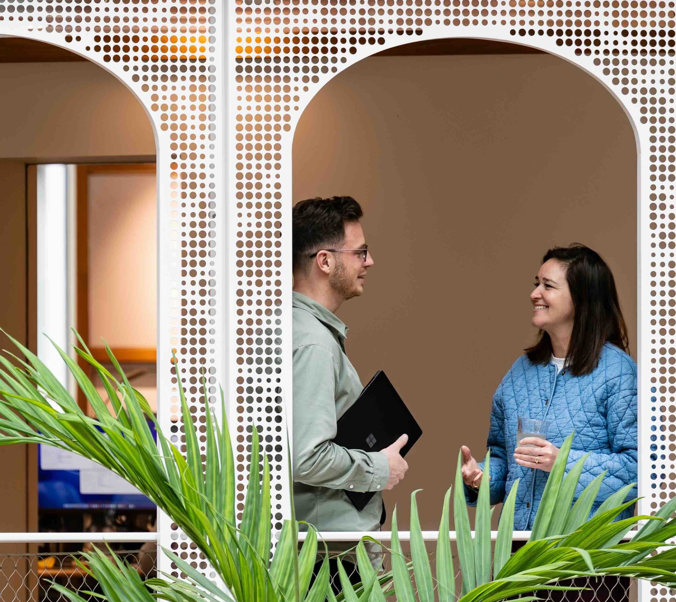 Hoofd Marketing en Partners VriendenLoterij Charlotte Regout staat met een collega te praten. Ze staan in het kantoor van de VriendenLoterij bij gebogen openingen in de muur. De man houdt een laptop vast en Charlotte een kop thee vast. Groene bladeren zijn te zien op de voorgrond.
