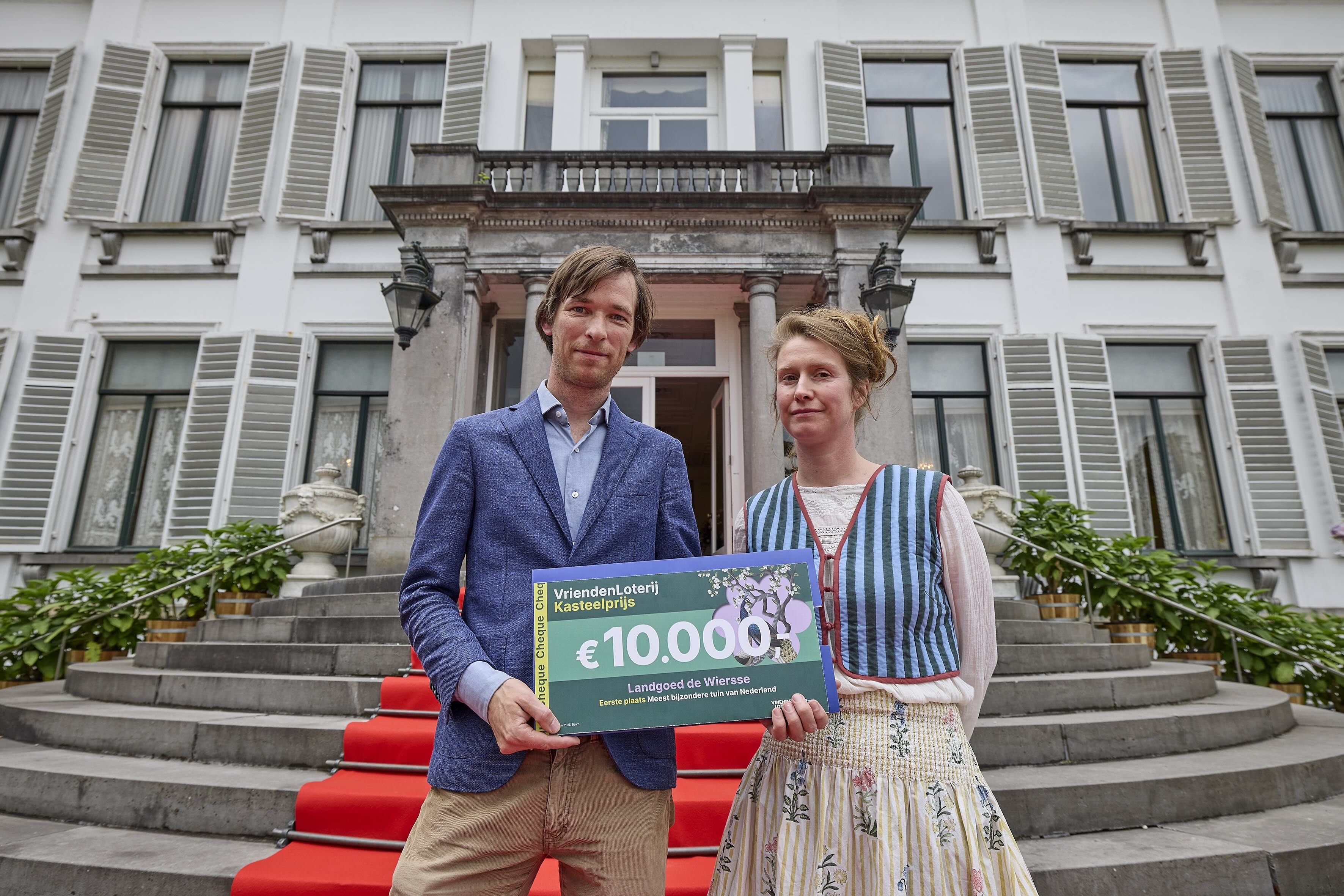 Twee winnaars van de Kasteelprijs staan voor de buitentrap met rode loper voor een kasteel met witte opengeslagen houten luiken en houden een grote cheque van €10.000 vast.