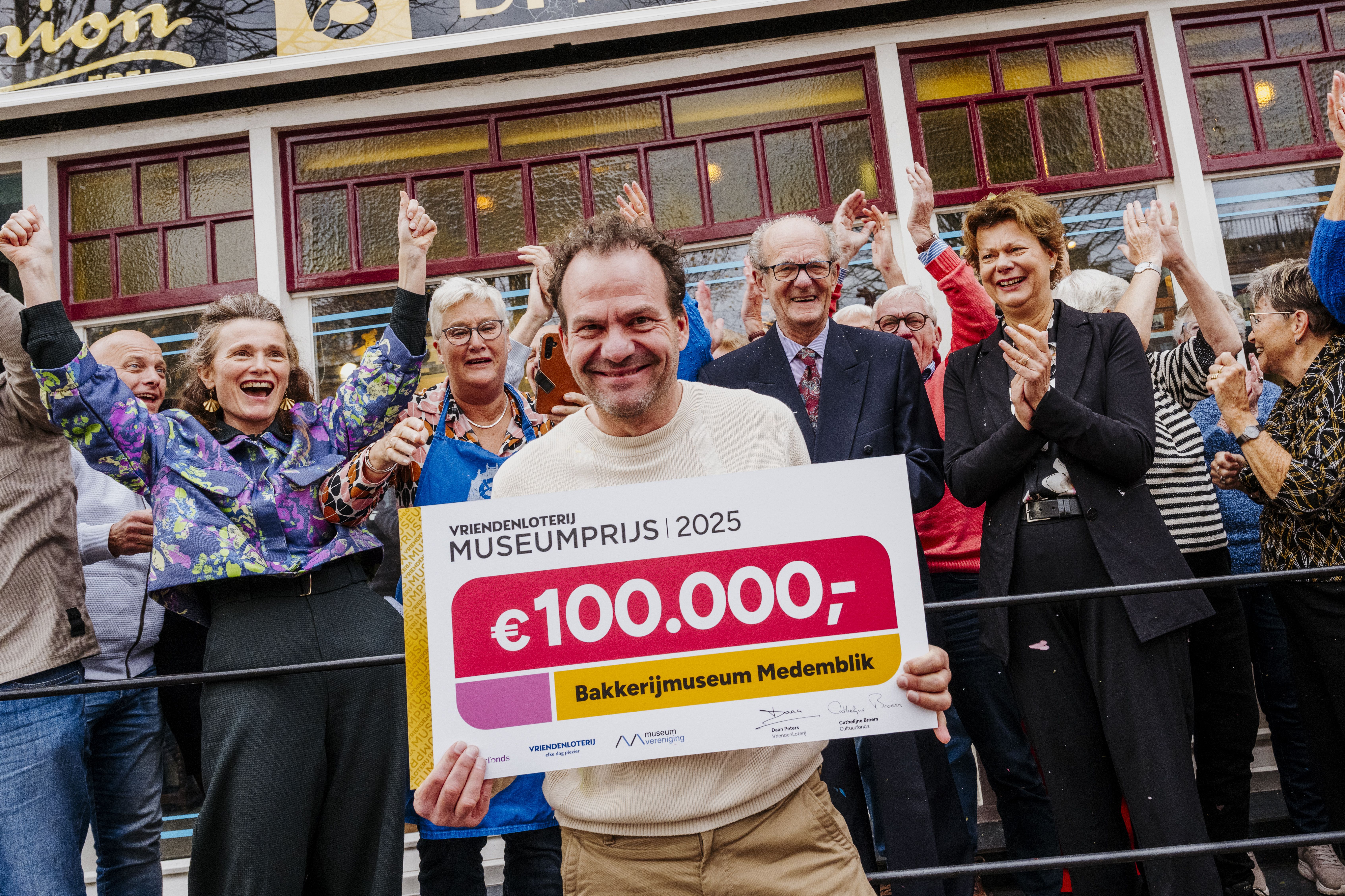 Vier mensen staan op een klassieke witte ophaalbrug en houden een grote cheque van de VriendenLoterij Molenprijs vast ter waarde van €55.060. Eén persoon heeft een bos bloemen.